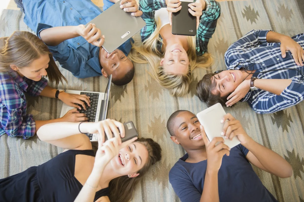 This is a photo of a bunch of kids laying down on the floor and instead of playing, they are on their devices. 